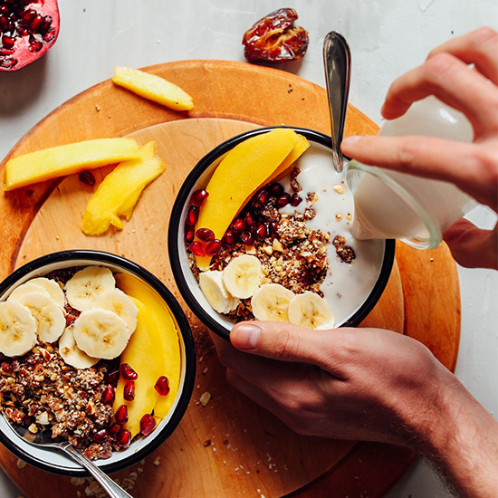 Pouring dairy-free milk into a bowl of Rawnola topped with fresh fruit