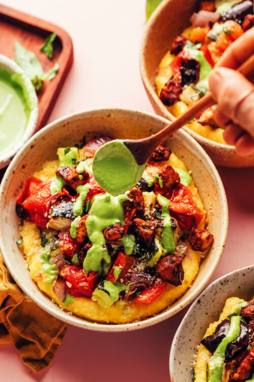 Drizzling pesto sauce onto a bowl of polenta with sheet pan ratatouille and tofu