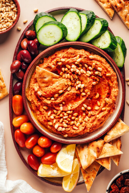 Bowl of sun-dried tomato hummus surrounded by sliced veggies and pita bread for dipping