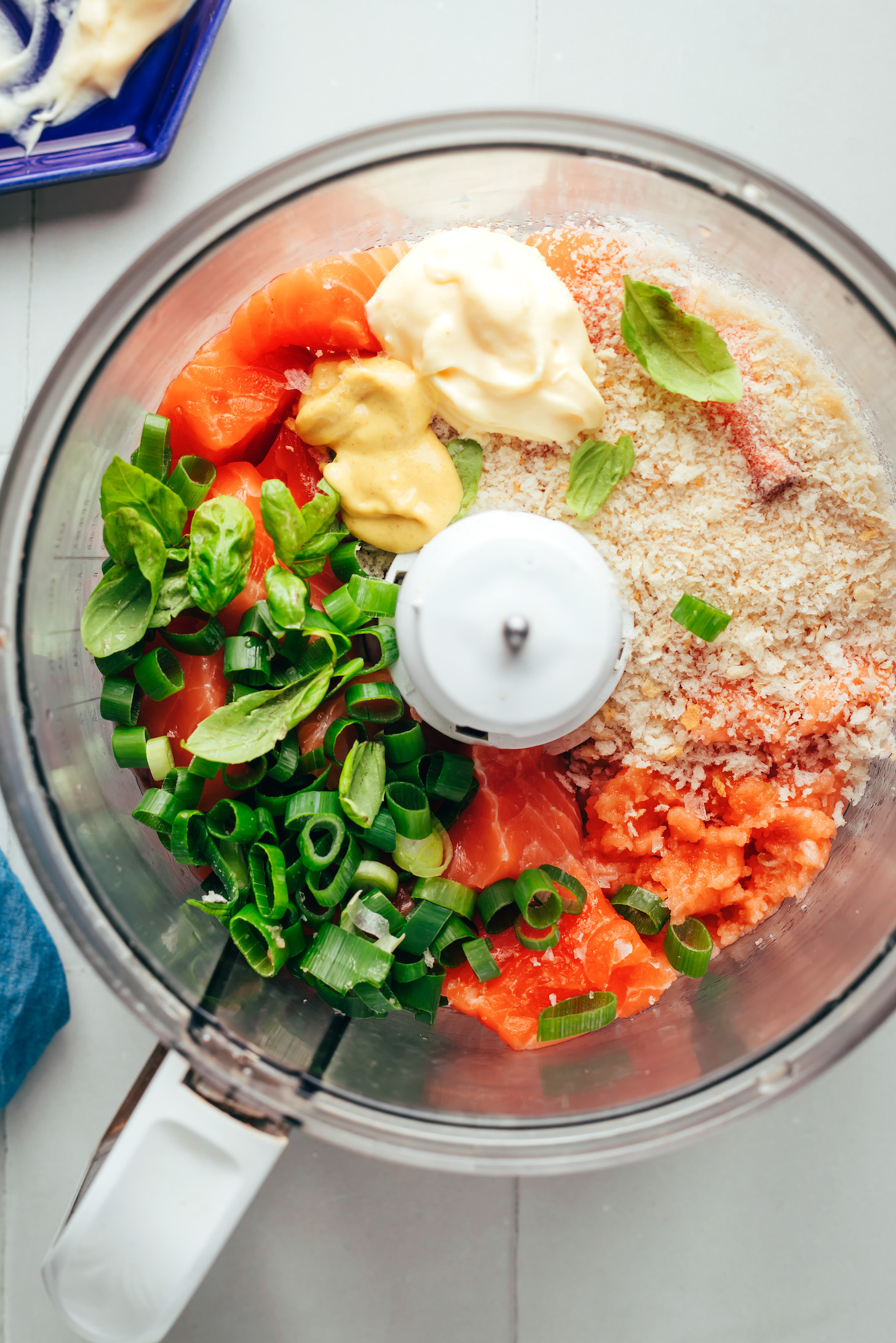 Food processor with cubed salmon, sliced green onion, fresh basil, mustard, mayonnaise, and breadcrumbs