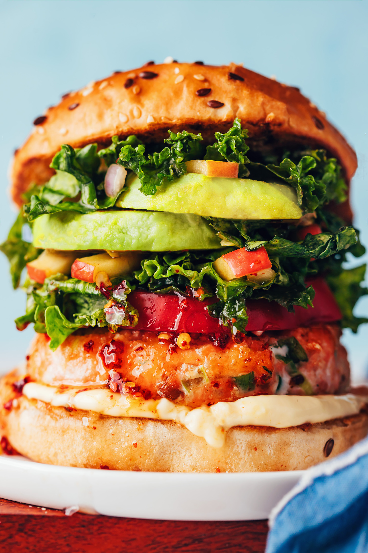 Close up photo of a salmon burger on a plate with mayonnaise, sweet chili sauce, tomato, kale apple slaw, and avocado